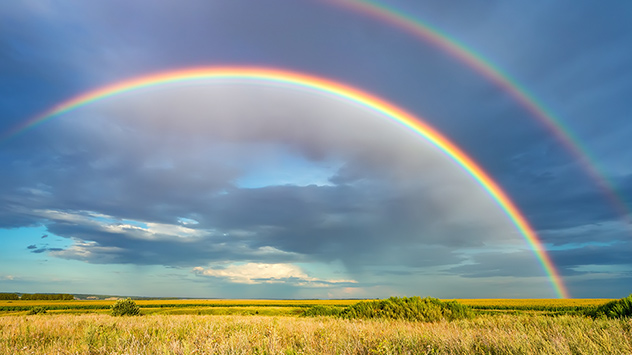Doppelter Regenbogen über Feldern vor wolkigem Himmel.