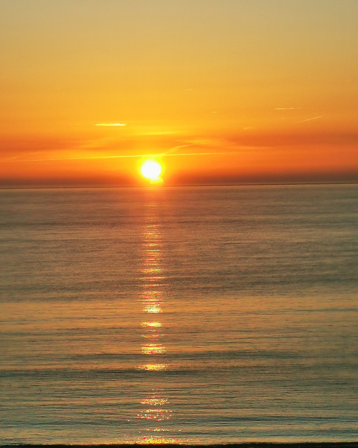 Bright sun setting over calm ocean with a deep golden sky, long shimmering reflection stretching across the water toward the shore.