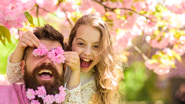 Mädchen hält einem Mann rosa Blüten vors Gesicht, beide lachen in frühlingshafter Umgebung mit blühenden Bäumen.