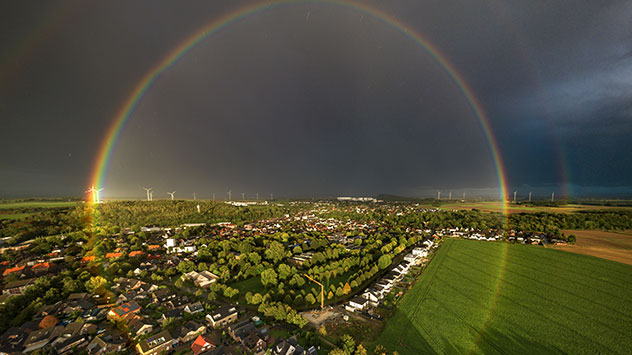 Doppelter Regenbogen über Feldern und Dorf mit dunklen Wolken im Hintergrund.