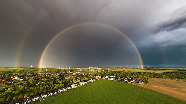 Doppelter Regenbogen über Feldern und Dorf mit dunklen Wolken im Hintergrund.