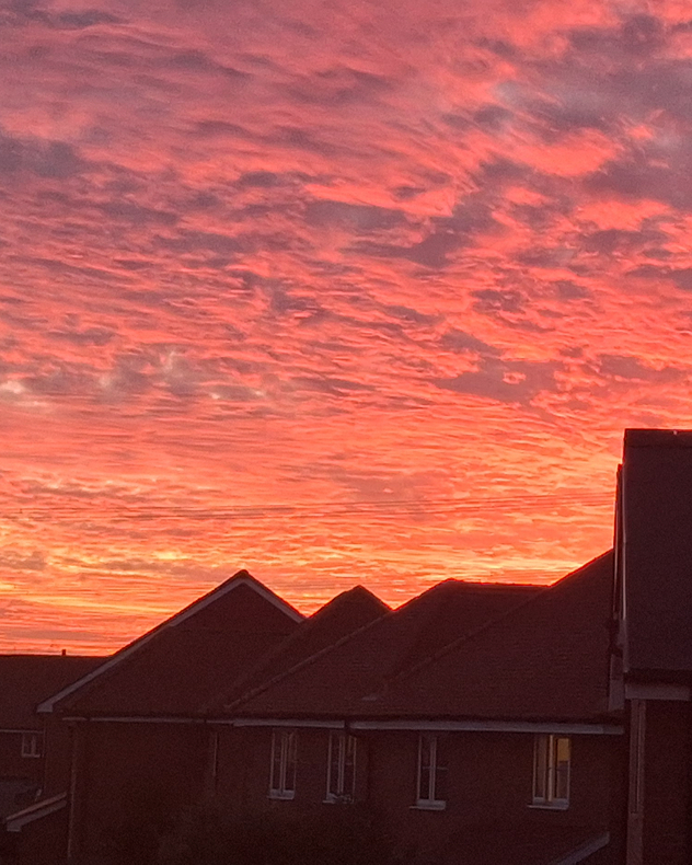 Intense red and orange sunset illuminates rippled clouds above dark silhouetted rooftops in a residential neighbourhood.