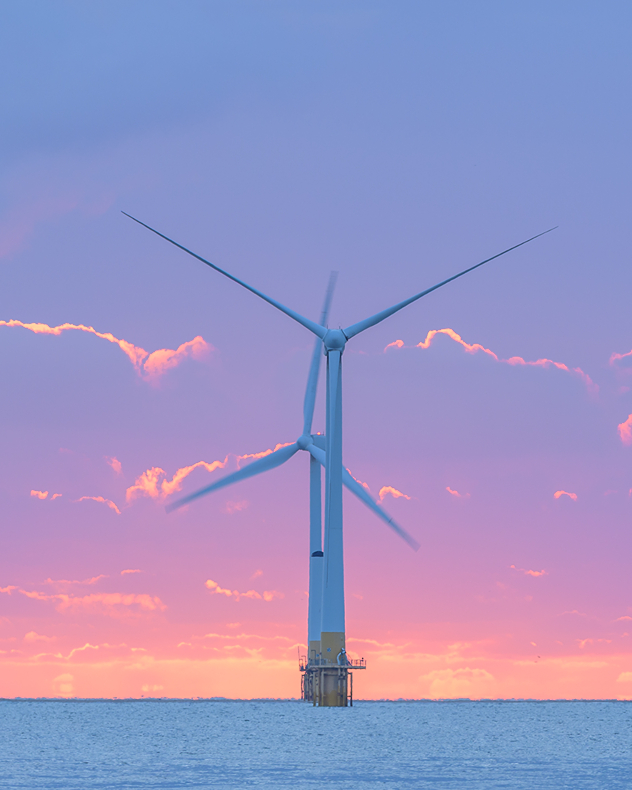 Two offshore wind turbines rise from calm blue water beneath a pink-purple sunset sky with glowing cloud edges.