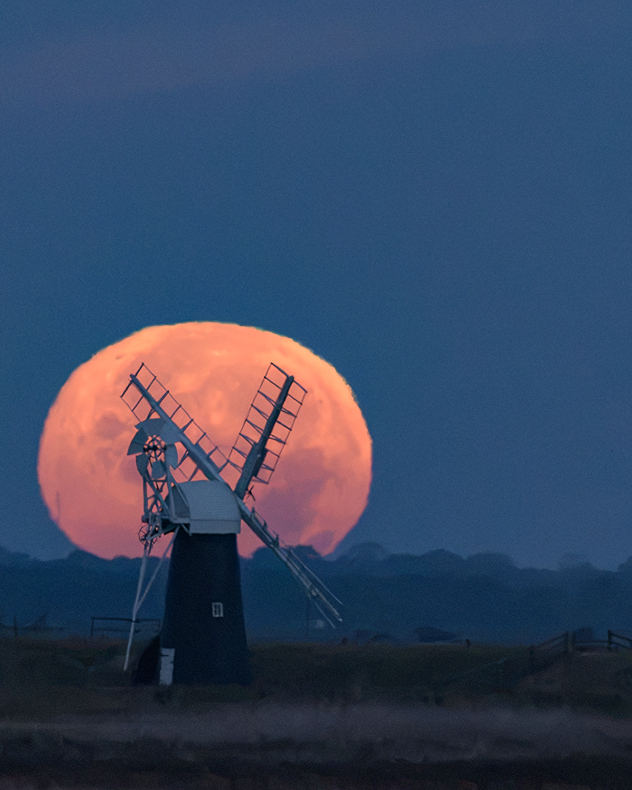 Large orange full moon rises behind a silhouetted traditional windmill beneath a deep blue twilight sky.