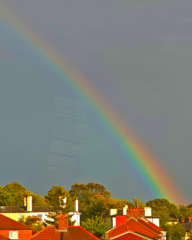 Bright rainbow arcs across a dark grey sky above sunlit red-roofed houses and autumn trees, with vivid red, yellow, green, and blue bands.