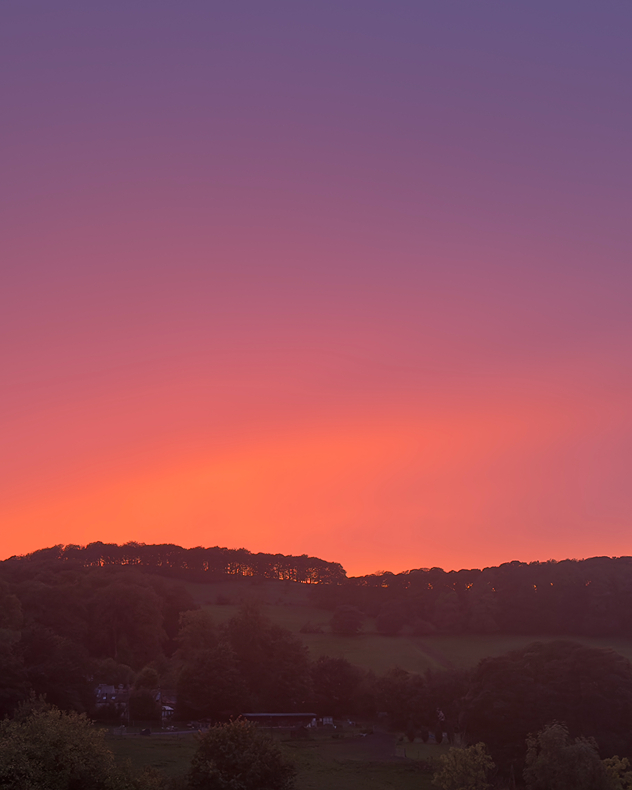 Vivid orange, pink, and purple sunset glows above dark silhouetted wooded hills and a quiet rural valley.