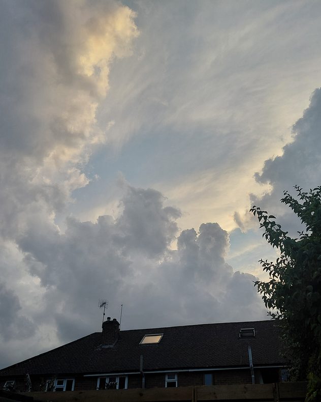 Towering gray cumulus clouds build above rooftops and trees, with pale sunlight illuminating cloud edges and wispy layers higher in the sky.