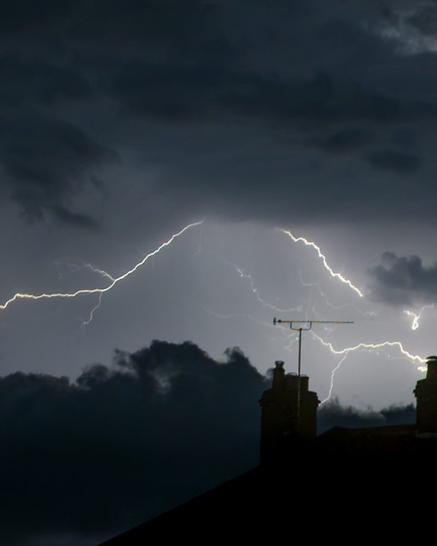 Multiple lightning bolts illuminate dark storm clouds above silhouetted rooftops, chimneys and a television aerial.