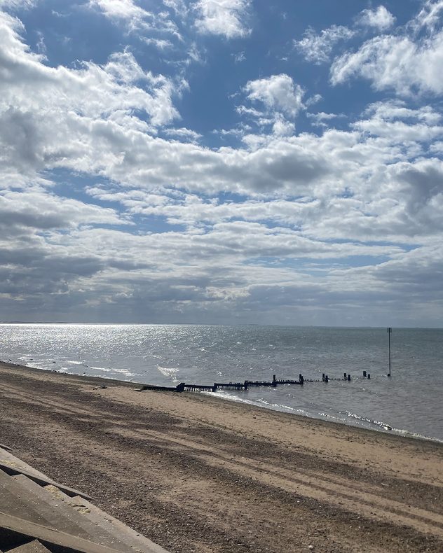 Quiet sandy beach beside a calm sea under broken cumulus clouds, with sunlight shimmering on the water and wooden groynes extending from the shore.