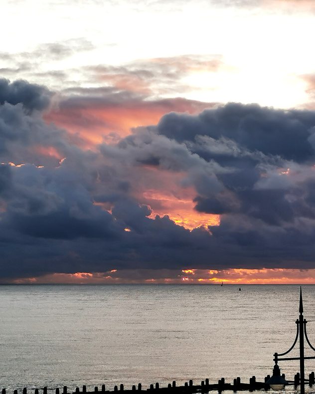 Dark layered storm clouds over a calm sea at sunset, with vivid orange-pink light breaking through gaps near the horizon and silhouetted seafront structures below.