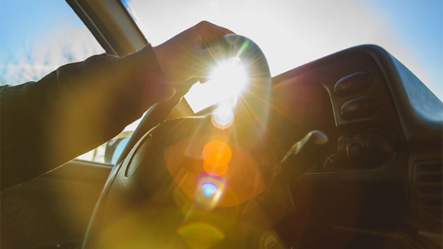Sonne blendet durch die Frontscheibe eines Autos, während eine Person das Lenkrad hält – starke Lichtreflexionen erschweren die Sicht beim Fahren.