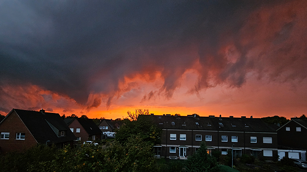 Dunkle, tief hängende Wolken ziehen über Häuserdächer. Am Horizont leuchtet der Himmel intensiv orange und rot.