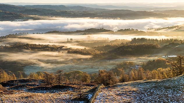 Hügelige Landschaft im Morgenlicht mit Bodennebel über Feldern und Wäldern. Die ersten Sonnenstrahlen brechen durch – vereinzelt liegt Raureif oder Schnee, was auf einen klaren und kalten Herbst- oder Wintermorgen hindeutet.