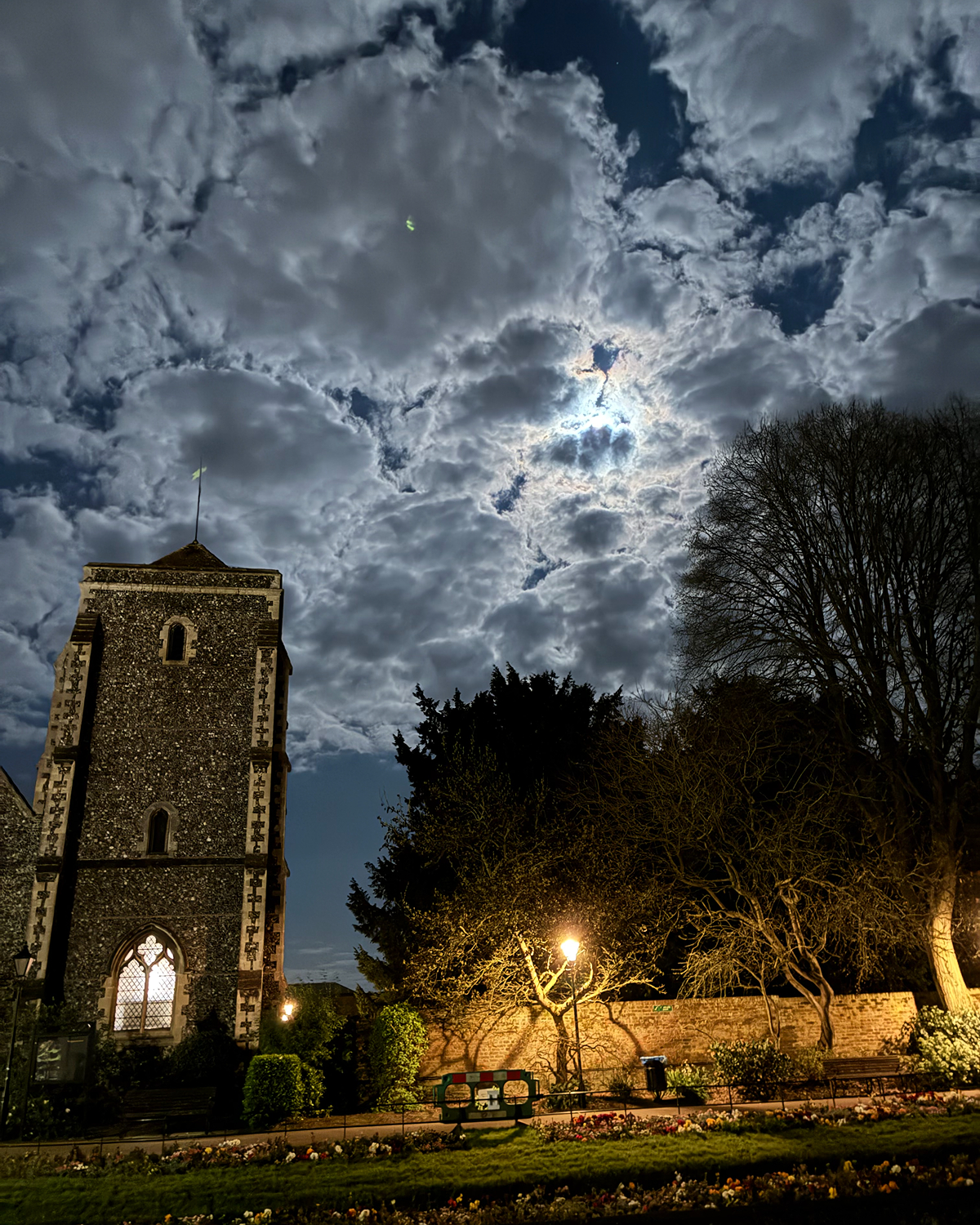 Historic stone church tower in Canterbury at night beneath dramatic moonlit clouds, with bright moonlight breaking through and trees and a lit streetlamp in the foreground.