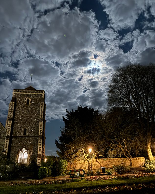 Historic stone church tower in Canterbury at night beneath dramatic moonlit clouds, with bright moonlight breaking through and trees and a lit streetlamp in the foreground.
