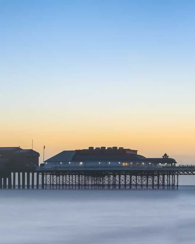 Cromer Pier in Norfolk at dawn with Pavilion Theatre silhouetted against a soft orange-blue sky, calm sea blurred in long exposure beneath the structure.