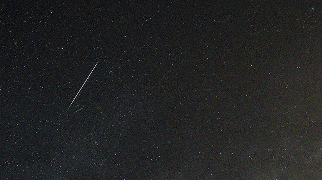Un brillante rayo de meteoro que atraviesa en diagonal un cielo nocturno repleto de estrellas, dejando una estela luminosa sobre un fondo oscuro salpicado de estrellas.
