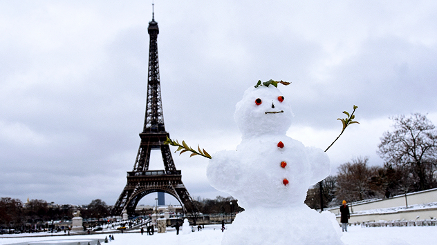 Előtérben egy hóember áll, ágakkal a karjai helyén. A háttérben az Eiffel-torony látható téli időben. A talaj és a környezet hóval borított.