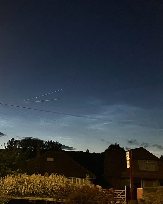 Electric-blue noctilucent clouds glowing above silhouetted houses and trees after sunset, with faint aircraft contrails and overhead utility wires crossing the deep twilight sky.