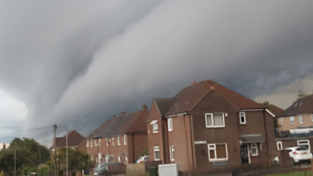 Shelf cloud looming over rows of brick houses, with the dark, dense cloud formation stretching horizontally across the sky, signalling an approaching storm.
