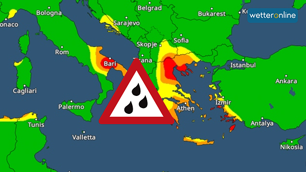 Mapa meteorológico con zonas de lluvias intensas marcadas en colores sobre Grecia y el sur de Italia. Las zonas rojas indican una alta intensidad de precipitaciones. El símbolo de alerta en el centro indica condiciones meteorológicas adversas.