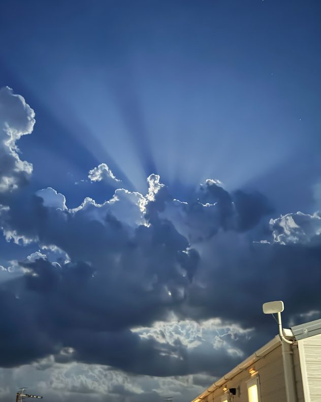 Dramatic crepuscular rays streaming from behind towering dark storm clouds, with silver-lined cloud edges against a deep blue sky above rooftops, creating a striking evening light display.