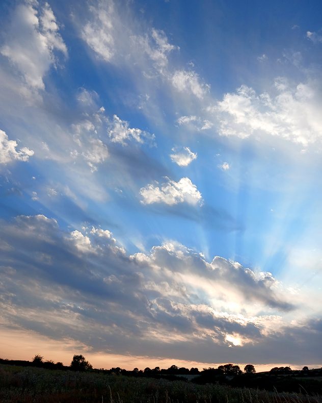 Dramatic crepuscular rays radiating through scattered cumulus clouds at sunset, with blue sky, sunlit cloud edges, and silhouetted fields and trees along the horizon.