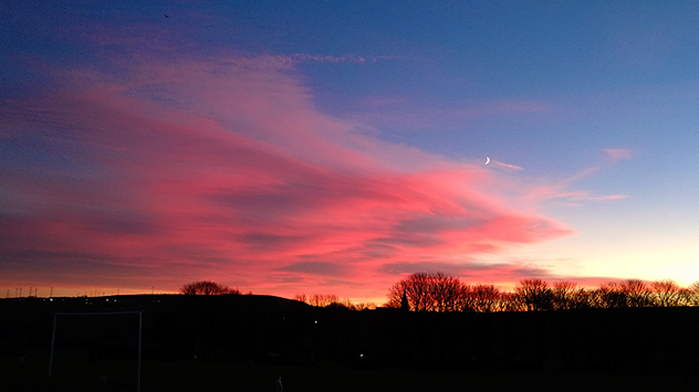 Waxing crescent Moon above vivid pink and magenta sunset clouds, with silhouetted trees along the horizon beneath a deepening blue twilight sky.