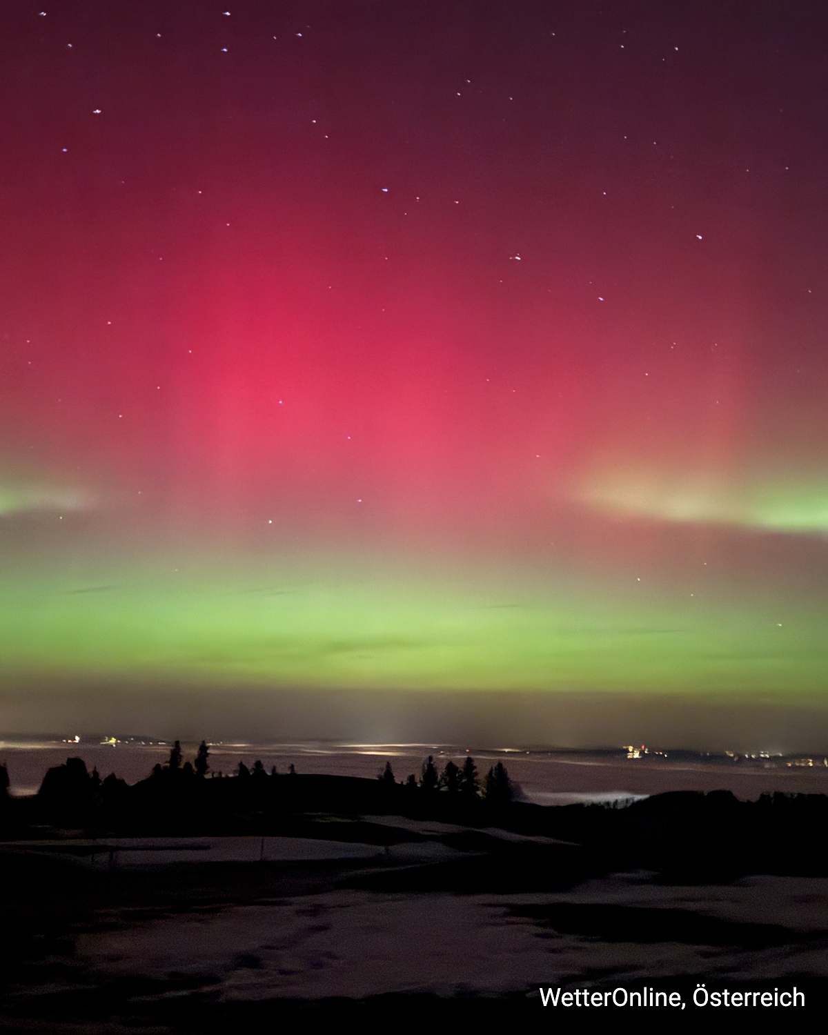 Green and red auroras over a snow-covered landscape in Austria. Starry sky with a glowing horizon.
