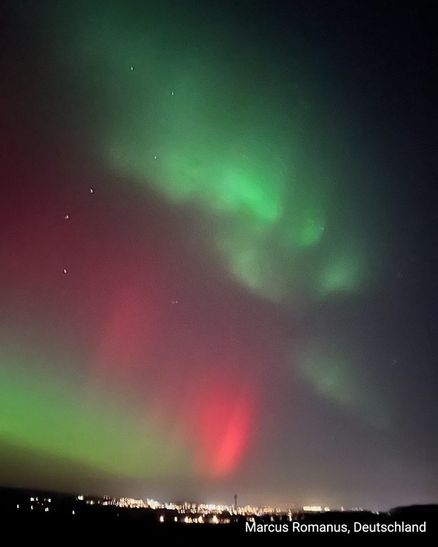 Green and red auroras over a German city at night. City lights visible on the horizon.