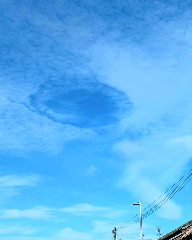 Oval-shaped fallstreak hole opens in a layer of small white clouds against a bright blue sky above rooftops and power lines.