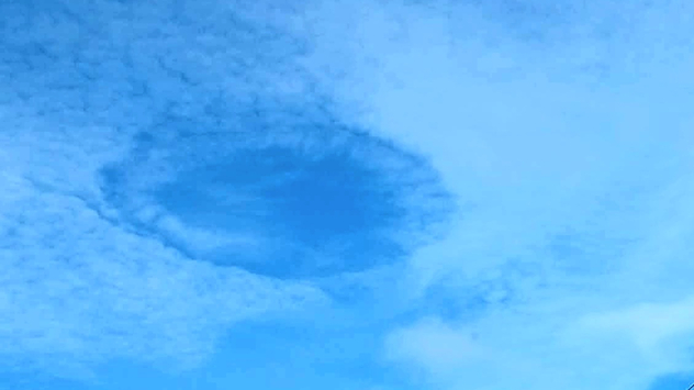 Oval-shaped fallstreak hole opens in a layer of small white clouds against a bright blue sky above rooftops and power lines.