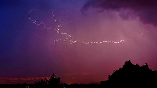 Heller, verzweigter Blitz in einem violetten Nachthimmel über der Stadt, dunkle Bäume und Dächer im Vordergrund.