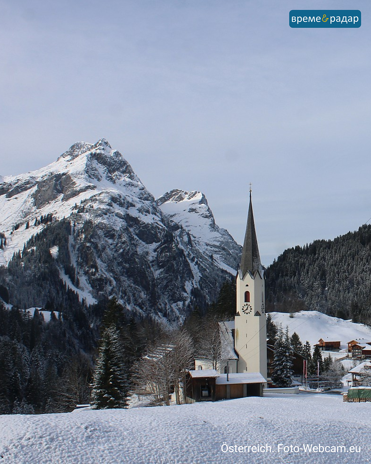 Заснежена църква в австрийско алпийско селище с високи планини на заден план.
