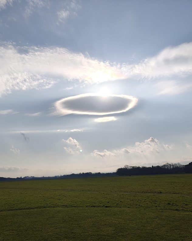 Bright sun behind a smooth lenticular cloud forming a glowing halo over an open green field and distant trees.