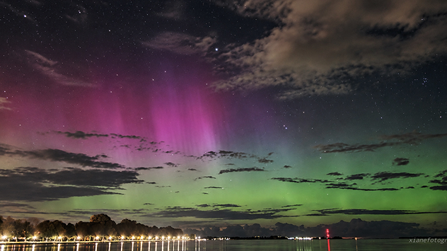 Rosa und grüne Polarlichter leuchten über Stralsund am sternenklaren Nachthimmel. Einzelne Wolken ziehen durch das Bild. Im Vordergrund ist eine dunkle Wasserfläche zu erkennen