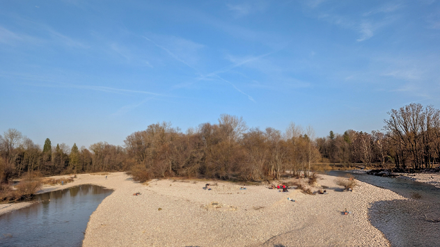 Fluss mit niedrigem Wasserstand und großem, trockenem Kiesufer. Menschen sitzen auf dem freigelegten Boden. Symbol für regenarmen März in Europa.