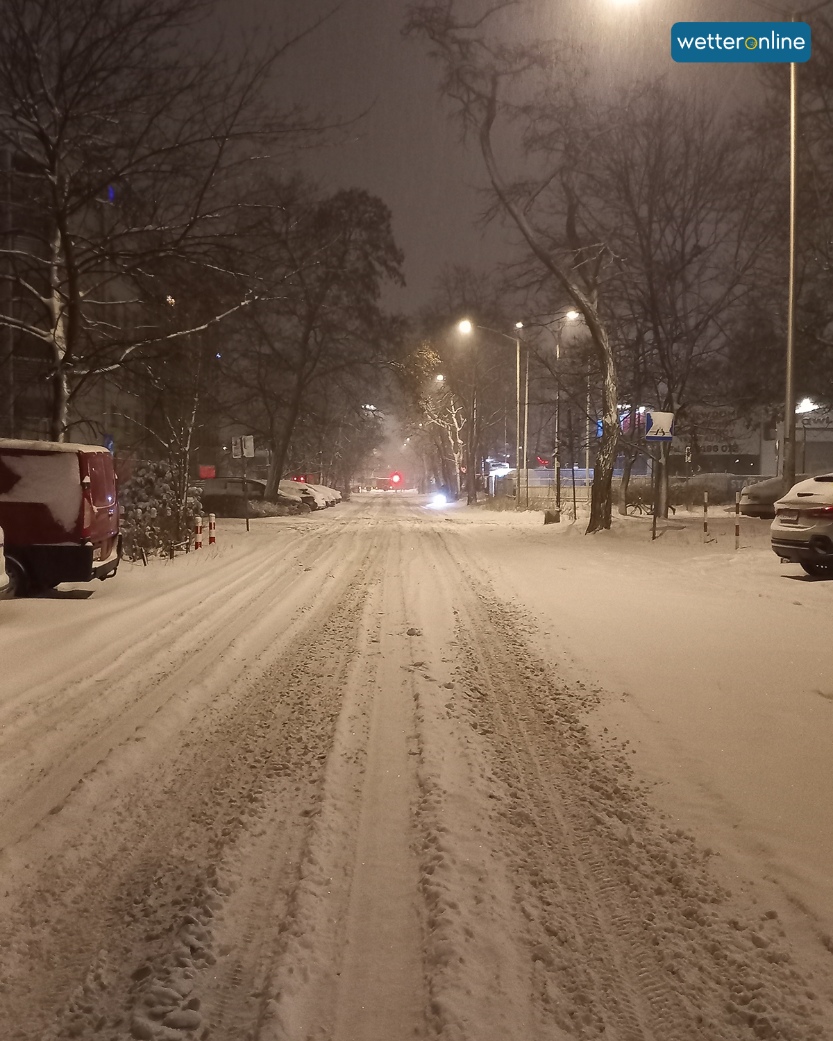 Snow-covered road with tyre tracks, illuminated by street lamps at night.