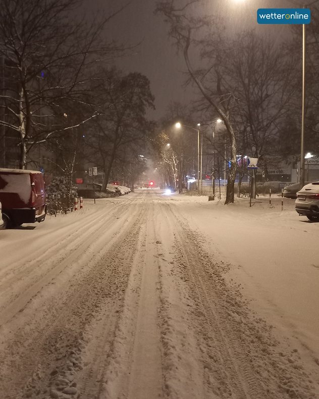 Snow-covered road with tyre tracks, illuminated by street lamps at night.