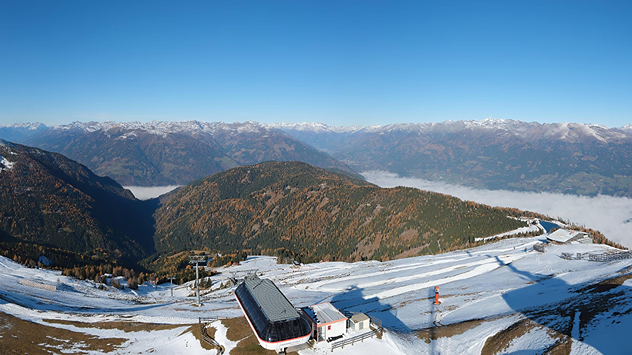 Das Bild zeigt eine Berglandschaft mit leicht verschneiten Hängen und einer Seilbahnstation im Vordergrund. Im Tal liegt Nebel, während im Hintergrund die Alpen unter klarem, blauem Himmel zu sehen sind.