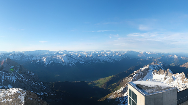 Das Bild zeigt ein weites Alpenpanorama mit schneebedeckten Gipfeln unter blauem Himmel. Im Vordergrund steht ein modernes Gebäude am Berggipfel mit Blick auf das sonnige Tal.