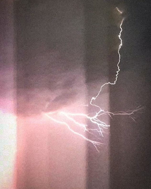 Bright branching lightning bolt illuminating the night sky beside a building, with multiple forked channels spreading across dark storm clouds during an active thunderstorm.