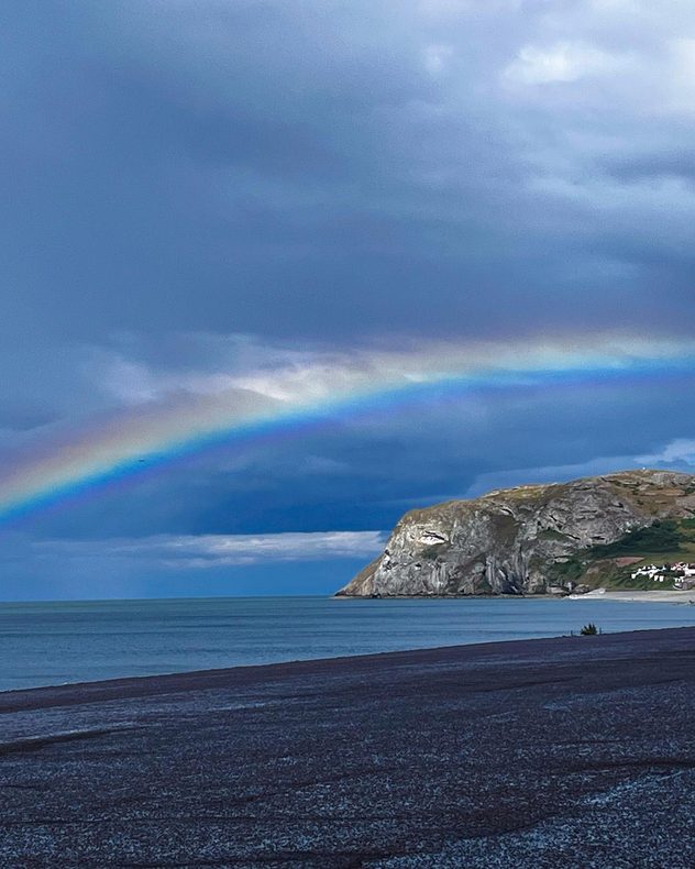 Bright rainbow arching over a calm sea beside white coastal cliffs beneath dark storm clouds, with a pebble beach and shoreline in the foreground.