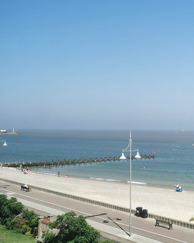 Sunny pebble beach with calm blue sea, wooden groynes, and a seaside promenade beneath a hazy blue sky, with a few people relaxing and swimming.