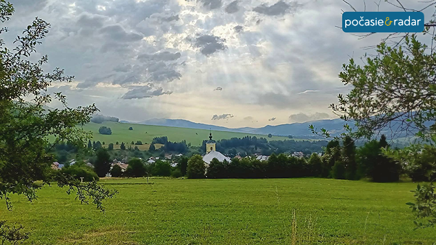 Slnečné lúče prenikajúce cez oblačnosť nad zelenou krajinou s kostolom a kopcami v pozadí