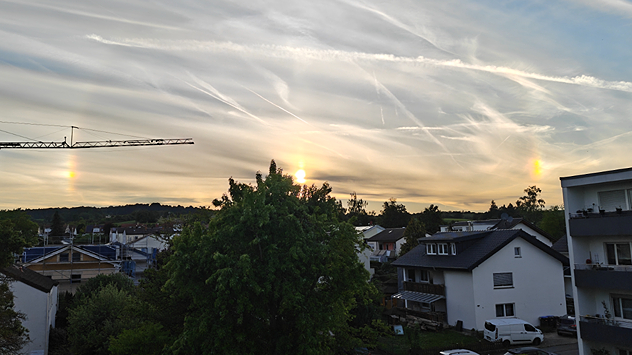 Abendhimmel mit Schleierwolken und sichtbaren Nebensonnen über einer Wohnsiedlung.