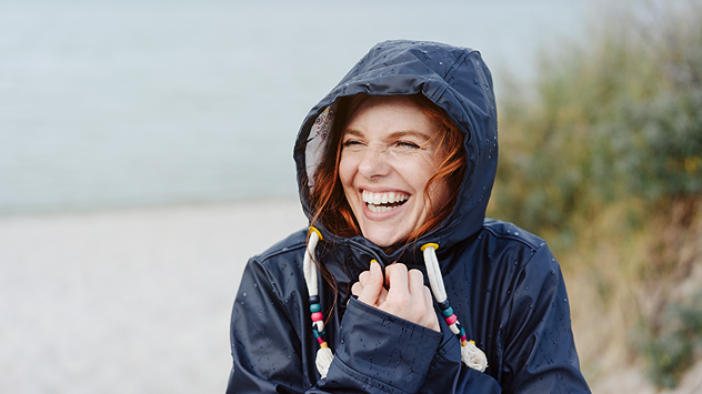 Lachende Frau mit roten Haaren trägt eine dunkle Regenjacke mit Kapuze am Strand. Tropfen auf der Jacke deuten auf nasses, windiges Wetter hin.