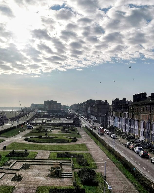 Elevated view of a coastal town seafront with a formal park and pathways in the foreground, terraced buildings lining the road to the right, light traffic, birds in the sky, and broken cloud with sunlight over the sea in the distance.