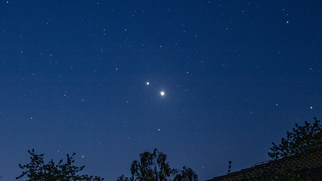 Ciel nocturne bleu avec de nombreuses étoiles. Deux planètes particulièrement lumineuses brillent au-dessus des silhouettes des arbres et des toits.