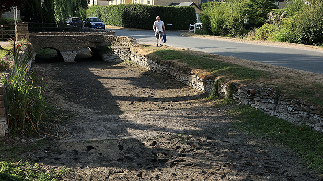 Dry section of the River Thames beneath a small stone bridge, with the riverbed exposed and cracked after prolonged, dry weather, while a person walks along the roadside above.